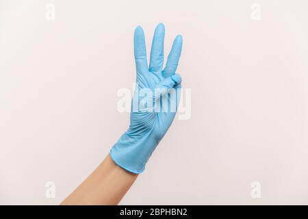 Vue latérale du profil gros plan de la main humaine avec des gants chirurgicaux bleus, portant le numéro 3 trois avec les mains. Vue en studio, à l'intérieur, isolée sur fond gris. Banque D'Images