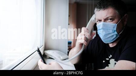 Un homme dans un masque médical se trouve devant une fenêtre et parle sur un téléphone fixe, tenant un smartphone dans la main. Il s'inquiète de la santé familiale duri Banque D'Images