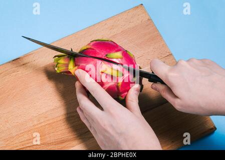 Gros plan des mains d'une femme coupant un fruit pitaya mûr avec un couteau sur une planche en bois. Pattaya est coupé en deux parties. Vue du dessus. Banque D'Images