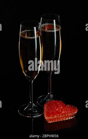 Deux verres de champagne et caviar rouge sur soucoupe en forme de coeur debout sur fond noir. Banque D'Images