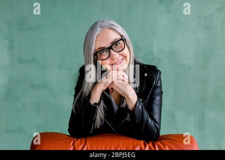 Studio portrait de charmante femme moderne senior en lunettes, avec de longs cheveux gris droits, portant une veste en cuir noir tendance, qui pose sur Banque D'Images