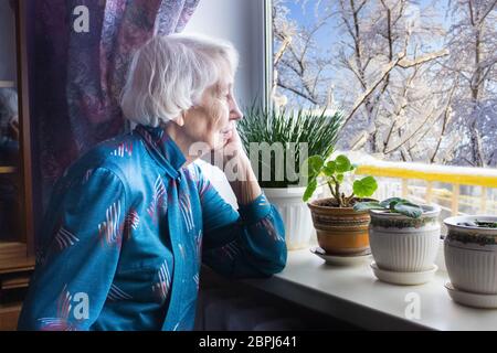 Vieille femme seule assise près de la fenêtre dans sa maison et regardant et rêvant Banque D'Images