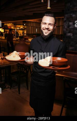 Serveur à barbe gai avec soupe de tomates riche, bœuf, riz. Un serveur éclairé en vêtements noirs, avec une barbe et une soupe de tomates Banque D'Images