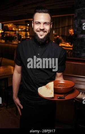 Serveur à barbe gai avec soupe de tomates riche, bœuf, riz. Un serveur éclairé en vêtements noirs, avec une barbe et une soupe de tomates Banque D'Images