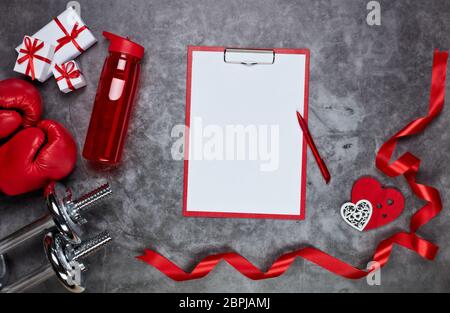 Haltères, gants de boxe, boîtes-cadeaux, coeurs rouges, bouteille d'eau et tablette avec feuille vierge sur fond gris.vue du dessus. Carte de Saint-Valentin. Fern Banque D'Images