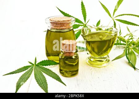 L'huile de chanvre dans deux pots en verre et saucière, feuilles et les tiges du cannabis sur l'arrière-plan de la lumière des planches Banque D'Images