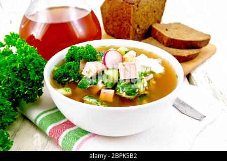 Soupe froide d'okroshka, pomme de terre, saucisses, oeufs, concombres, radis verts et kvass dans un bol blanc sur une serviette, pain et verre à verre sur l'arrière-plan Banque D'Images