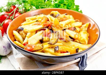 Pâtes penne à l'aubergine et les tomates dans un bol sur une serviette, une fourchette, l'ail et au persil sur une planche en bois historique Banque D'Images