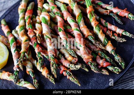 Apéritif facile asperges enveloppées de bacon d'érable grillées à l'ail et au sel de mer servies sur une assiette noire avec citron sur un fond en bois ancien, vue de dessus Banque D'Images