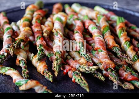 Apéritif sain asperges enveloppées de bacon grillées à l'ail et au sel de mer servies sur une assiette noire avec citron sur un fond en bois ancien, vue sur le dessus, c Banque D'Images