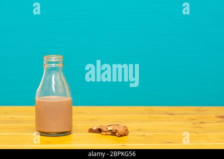 Un tiers de pinte de lait en verre bouteille à moitié plein avec du chocolat milkshake et demi-biscuit aux brisures de chocolat mangé, sur une table de bois contre un teal backgroun Banque D'Images