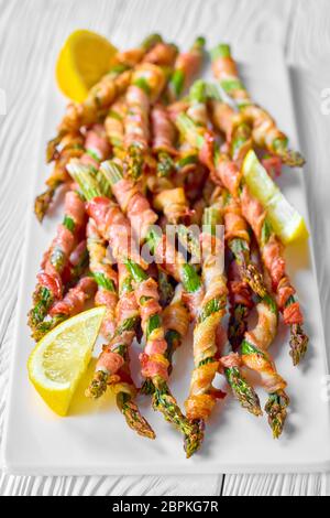 Céto régime bacon asperges enveloppées de bacon rôti à l'ail et sel de mer servi sur une assiette blanche avec citron sur fond de bois blanc avec l'eau Banque D'Images