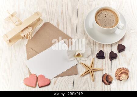 Tasse de café, biscuits et chocolats sous forme de coeur, coquillages, avion et feuille vierge avec enveloppe sur fond de bois clair. Haut vi Banque D'Images