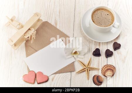 Tasse de café, biscuits et chocolats sous forme de coeur, coquillages, avion et feuille vierge avec enveloppe sur fond de bois clair. Haut vi Banque D'Images