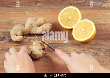 Les mains des femmes coupées en morceaux de racine de gingembre pour une boisson faite de citron frais et de gingembre sur fond de bois brun. Boisson artisanale. Banque D'Images