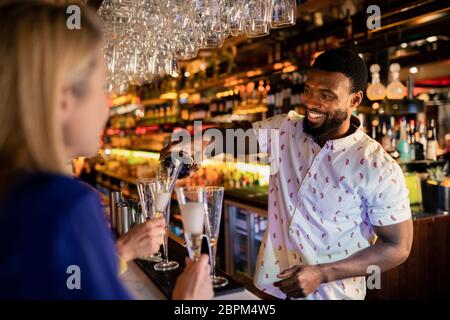 Barman de la mise à niveau de maturité avec un verre de prosecco de womans. Elle est assise sur le comptoir du bar avec ses amis. Banque D'Images