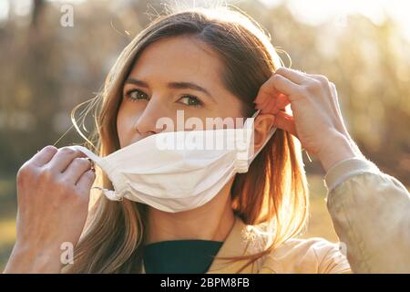 Jeune femme mettant sur le coton blanc fait maison virus nez bouche masque, parc de bokeh flou en arrière-plan. Prévention des épidémies de coronavirus covid-19 Banque D'Images