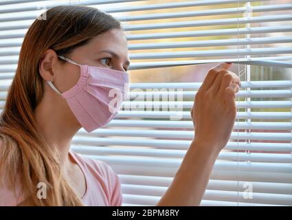 Jeune femme en rose fait maison de coton virus masque, regardant à travers les stores de fenêtre dehors. Mettre en quarantaine ou rester à la maison pendant le coronavirus covid-19 ou Banque D'Images