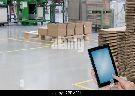 Hall de production vide avec des boîtes en papier plié sur des palettes en bois et les mains tenant tablette avec écran blanc prêt pour votre texte. Banque D'Images