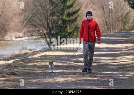 Jeune homme en veste rouge, bonnet et lunettes de soleil, portant le masque de visage de virus fait maison, marche chien sur la route de campagne. Les masques sont obligatoires en dehors de la maison pendant Banque D'Images