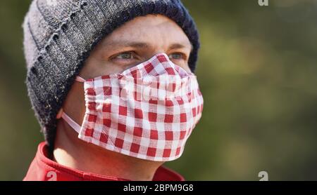 Jeune homme en bonnet beanie chaud portant un coton fait maison visage bouche nez masque de virus, parc ensoleillé flou en arrière-plan, peut être utilisé pendant le coronavirus cov Banque D'Images