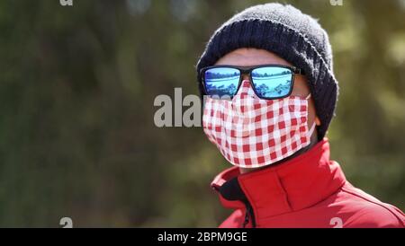 Homme portant un bonnet beanie, des lunettes de soleil bleues brillantes et du coton blanc rouge fait maison nez bouche visage masque de virus, gros plan portrait de détail, parc flou arbres b Banque D'Images