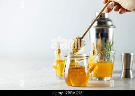 La main d'une femme tient une cuillère pour le miel sur le pot devant deux verres de cocktail de bourbon de miel avec du sirop de romarin ou du whisky maison aigre avec de l'orange Banque D'Images