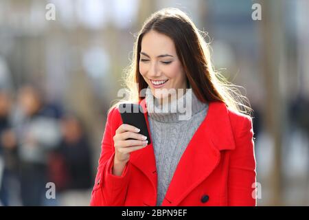 Une femme heureuse portant un manteau rouge marchant et utilisant un smartphone dans la rue en hiver Banque D'Images