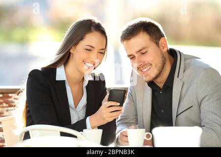 Deux cadres heureux regardant du contenu en ligne sur un téléphone portable assis sur une terrasse de restaurant Banque D'Images