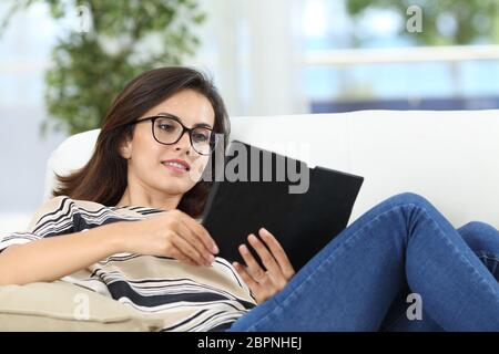 Femme heureuse de lire un livre assis sur un canapé dans la salle de séjour à la maison Banque D'Images