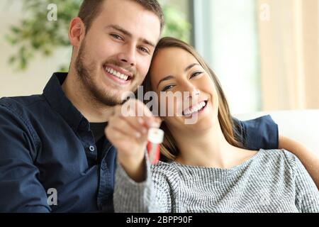 Heureux couple de propriétaires montrant des clés de la maison à la caméra assis sur un canapé dans la salle de séjour à la maison Banque D'Images