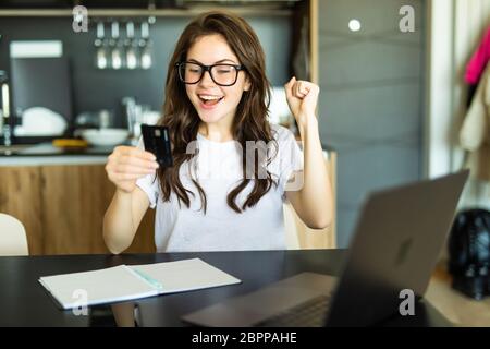 Image d'une belle jeune femme brunette émouvante et émouvante dans la cuisine à l'intérieur à la maison avec ordinateur portable tenant la carte de crédit. Banque D'Images