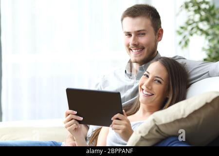 Couple holding a tablet posant assis sur un canapé à la maison et looking at camera Banque D'Images