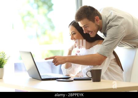 Vue latérale d'un couple heureux recherchant des informations en ligne sur un ordinateur portable sur une table à la maison ou dans une chambre d'hôtel avec une fenêtre en arrière-plan Banque D'Images