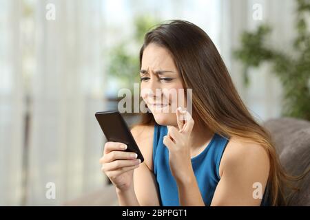 Fille d'espoir crossing fingers holding un smartphone dans l'attente de nouvelles assis sur un canapé dans la salle de séjour à la maison Banque D'Images