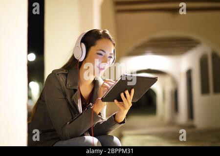 Relaxed woman wearing headphones regarder le support d'un comprimé le soir dans la rue Banque D'Images