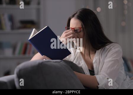 Dame portant des lunettes souffrant de fatigue visuelle de lire un livre assis sur un canapé à la maison dans la nuit Banque D'Images