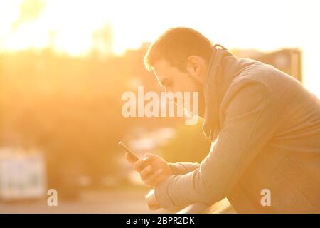 Vue latérale du portrait d'un homme à l'aide d'un téléphone intelligent en hiver dans une chambre balcon au coucher du soleil Banque D'Images