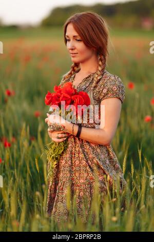 Portrait de la belle femme à tête rouge sur un terrain vert avec bouquet de coquelicots au coucher du soleil d'été Banque D'Images