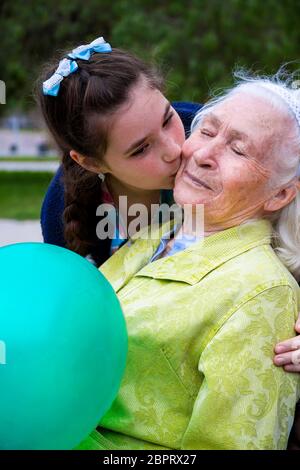 Pretty girl hugs et embrasse sa belle grand-mère aux cheveux gris sur la joue. Une agréable femme âgée est assise sur un banc de parc tenant un ballon et sm Banque D'Images