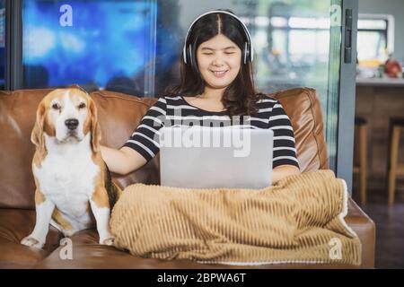 Femme d'affaires asiatique utilisant un ordinateur portable et un casque pour travailler à domicile dans une maison en salle par vidéoconférence, assis avec un chien, startups Banque D'Images