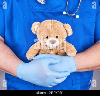 Médecin en uniforme bleu et des gants en latex tenant un ours en peluche brun, fond gris Banque D'Images