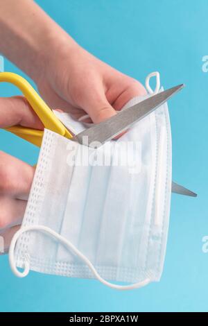 L'homme coupe un masque de protection médicale avec des ciseaux jaunes sur fond bleu. L'idée de conspiration et de incrédulité dans le danger du coronavirus Banque D'Images