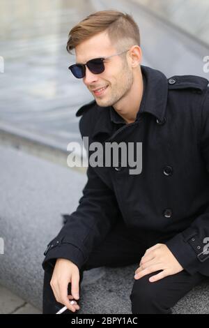 Jeune homme blond souriant portant une veste noire assis sur la Pyramide du Louvre et fumant. Banque D'Images