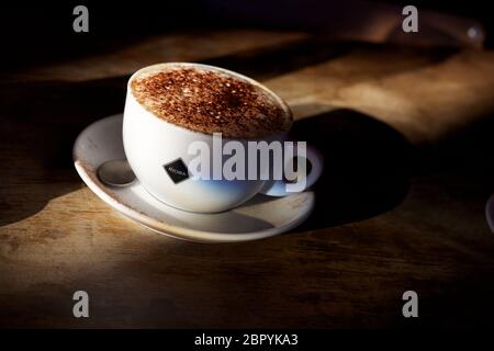 Un cappuccino dans une tasse blanche avec dépoussiérage de poudre de chocolat sur le dessus et dépoussiérage de poudre de chocolat sur la soucoupe et la table blanches, Banque D'Images