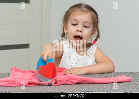 une jolie petite fille de 4 ans joue une femme au foyer, repassant les vêtements des enfants avec un fer à repasser Banque D'Images