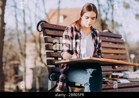 Jeune artiste dessine une image tout en étant assise sur un banc. Artiste peintre tient une tablette en bois pour la peinture et un pinceau dans sa main Banque D'Images