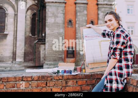 Une artiste femme peint une image tout en étant assise sur un mur de briques sur un fond d'architecture ancienne. Une fille passe des loisirs à dessiner des images dans une rue de ville Banque D'Images