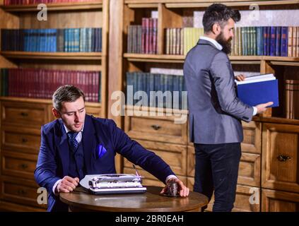 Auteur ou jeune auteur avec machine à écrire chercher de l'information pour un nouveau livre dans la bibliothèque avec un ami. Hommes en costume dans la bibliothèque avec des livres anciens avec des étagères sur fond. Concept d'artisanat des écrivains. Banque D'Images