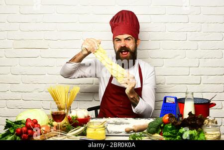 Chef fait de la pâte. Cuisinez avec un visage excités en uniforme bordeaux, assis près d'une table avec des légumes et des ustensiles de cuisine. Homme avec barbe étire la pâte à pétrir sur fond blanc. Concept de cuisine professionnelle Banque D'Images